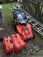 Full view of the Black & Decker lawn mower with attached leaf bag and two red gas cans in front on ground near fence and ladder.