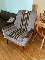 Front right angled view of the armchair showing fabric stripes, wooden armrests, and legs on hardwood floor, with other furniture partially visible.