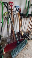 Collection of garden tools including shovels and forks standing against a wall, various handle colors and metal heads visible.
