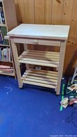 Full front view of the unfinished wood kitchen cart showing top, drawer, two slatted shelves, and wheels.