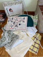 Photo showing assorted folded table linens including rectangular and round tablecloths, place mats, napkins, and a teal colored tea cozy.