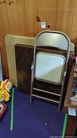 Metal folding chair with speckled cream cushioned seat, shown folded and leaning against two folding card tables.