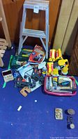 Lot arranged on blue carpet with wooden wall backdrop showing four yellow/red flashlights, small metal toolbox, multiple hand tools, hot glue guns, maintenance items, and a small silver folding ladder