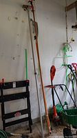 Three long tree trimming tools leaning against a wall in a garage including saw, rake, and limber. Handles are mostly wood, one with red operating mechanism at top.