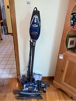 Full view of blue Shark Rocket Deluxe Pro vacuum standing upright with hose wrapped and multiple attachments and instruction manual on the floor, showing included items.