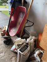 Photo showing the red wheelbarrow with visible dirt and wear inside, Black & Decker cordless trimmer standing against wheelbarrow, several bags of Sakrete and stones on the floor around it.