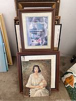 Lot of large framed artworks, showing a painting of a woman with a baby and another with an outdoor scene, along with stacked frames behind.