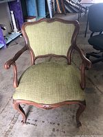 Front view of antique wooden armchair with green fabric upholstery, showing carved wooden frame and seat cushion.