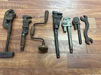 Full view of all eight antique hand tools on wooden floor showing pipe wrenches, hand drill, and spanner wrench