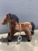 Side view of a brown plush ride-on horse toy with black mane and tail, fabric saddle, stirrups, and wooden handle on side