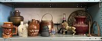 Shelf with various ceramic and pottery vases and jugs, brass horse figurine, vintage Pepsi bottle, brass decorative trivet, and rusty metal stirrups.