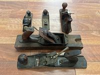 Five metal hand planers arranged on floor showing size and condition: oxidation, paint fading, wooden handles and knobs visible.