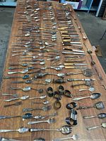 Overview of a large collection of mixed metal flatware spread on wooden table including spoons, forks, knives, serving pieces, and napkin rings needing polishing.