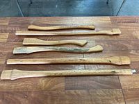 Six wooden axe handles laid side by side on a wooden table, showing variety in shape and size, visible scratches and wear.