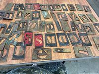 Metal stencil lot spread out on a wooden surface showing a variety of letters and numbers, some painted red, all with signs of age and wear.