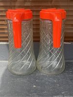 Two vintage Anchor Hocking Tang glass pitchers with orange plastic handles and lids, showing their embossed swirl pattern and slight cloudiness of the glass due to age.