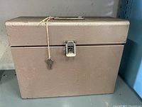 Front view of a rectangular vintage metal lock box in textured light brown with a silver latch and two keys tied to the latch with string.