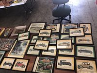 Overview photo showing a large number of framed vintage pictures on table including multiple classic car prints and some landscape art.