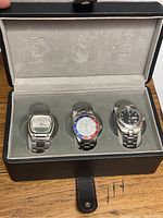 Full view of black leather watch box with gray velvet lining, holding three metal band watches.