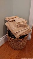 Photo showing two woven rattan baskets stacked with a pile of beige towels of various sizes on top, placed on hardwood floor against a wall.