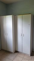 Two tall white storage cabinets side by side, showing their front with closed double doors and simple handles, positioned against a pale blue wall.