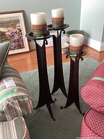 Three dark metal candle holders with tall legs, featuring large white and brown banded pillar candles, arranged on carpeted floor.