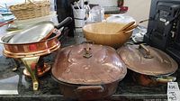 Overview photo showing two copper baking pans with lids and handles, a wooden bowl with salad servers, and a copper chafing dish with stand and burner on kitchen counter.