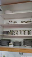 Four-shelf cabinet with plain white mixing bowl on top, sugar jar, spice containers, and gravy boat on second, small cups and mugs on third, plates and bowls on bottom.