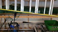 Photo showing two aluminum ladders placed horizontally on a wooden shelf. One is an extended straight ladder (extension ladder) and the other is a shorter folding ladder folded flat. Some cords and equipment are visible below the ladders.
