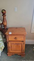 Frontal view of one wooden night stand with drawer and cabinet door next to a bed post.