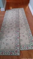 Two cream/off-white runners with floral border pattern, laid out on hardwood floor showing full length and design.