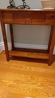 Full view of pine table against the wall, showing drawer with handle and lower shelf. Wooden floor beneath.