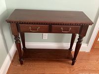 Full view of wooden table showing the top surface, two drawers with brass metal handles, turned legs, carved edge detail, and bottom shelf.