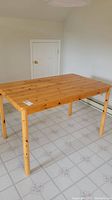 Rectangular wood table shown from an angle with view of floor and wall behind. The table has square legs and visible wood grain with knots.