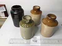 Four antique stoneware crocks on a white surface with measuring ruler and lot number tag. Crocs show varied glazes in brown, tan, green and dark brown colors.