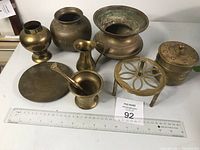 Eight assorted brass items displayed on a table with a measuring ruler, including vases, mortar and pestle, plate, trivet, and lidded container.