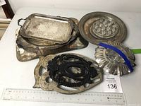 Top view showing a stack of various silver plated trays and metal trivets including an owl-shaped trivet and glass flower frogs.