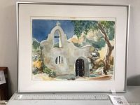 Frontal view of the framed watercolour artwork showing the church scene with tree on right, rocks left, and clear blue sky background.
