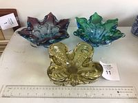 Three vintage heavy art glass ashtrays displayed on a table with a measuring ruler. Purple star-shaped, green star-shaped, and amber leaf-shaped ashtrays shown from top view.