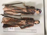 Top-down photo showing two vintage Indonesian puppets made of carved wood with fabric clothing, displayed with rulers for scale.