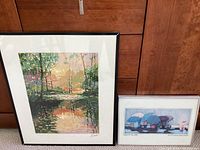 Two framed prints by Beth Hendrickson Logan. Left print depicts colorful reflections of trees on water with black frame. Right print shows whimsical characters with umbrellas and a pink frog in the rain, silver frame.
