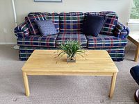 Front view of navy plaid couch with two throw pillows, blonde wood coffee table with small green plant décor on it in front of the couch, showing carpeted floor and adjacent furniture edge.