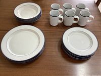 Full view of six smaller plates, five dinner plates, and six mugs with navy blue rims on a wooden table, illustrating the dish set for six minus one dinner plate.
