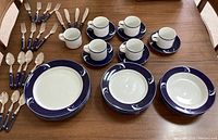 Full view of the dinnerware set consisting of stacked plates (dinner, small, salad bowls), cups, saucers, and flatware arranged on a wooden table.