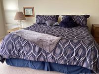 Full view of king-sized bed with peacock blue bedding, pillows, gray throw, lamp on side table to left.