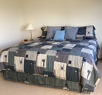 King sized bed with geometric patterned bedspread in shades of grey, blue, cream, and beige. Multiple pillows including grey and blue with tassels.