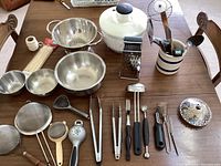 Full view of all kitchen items on wooden table: bowls, salad spinner, various utensils and strainers.