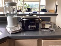 View of all three items together on kitchen counter, showing KitchenAid blender, black Crock-Pot slow cooker, and GE electric mixer with attachments.