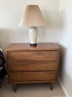 Front view of mid-century wooden dresser with three drawers and ceramic lamp on top.