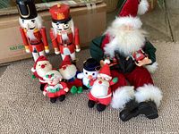 Photo showing two wooden nutcracker soldiers, multiple small Santa and snowman plush figures, and a large plush Santa holding toys, with a cardboard box in background
